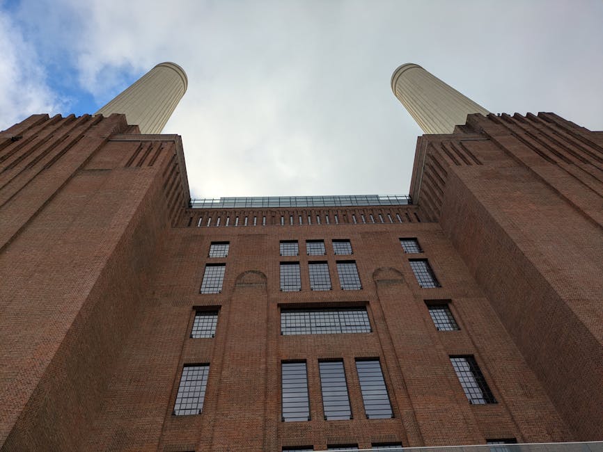 An upward-looking view of Battersea Power Station surrounded by modern glass and steel skyscrapers under a partly cloudy sky. The scene captures the contrast between the historic brickwork of the powe