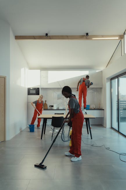 A woman in a grey t-shirt and red overalls is performing surface cleaning in a modern kitchen. She is using a mop or cloth to clean the floor next to a small blue bucket. The kitchen features white wa